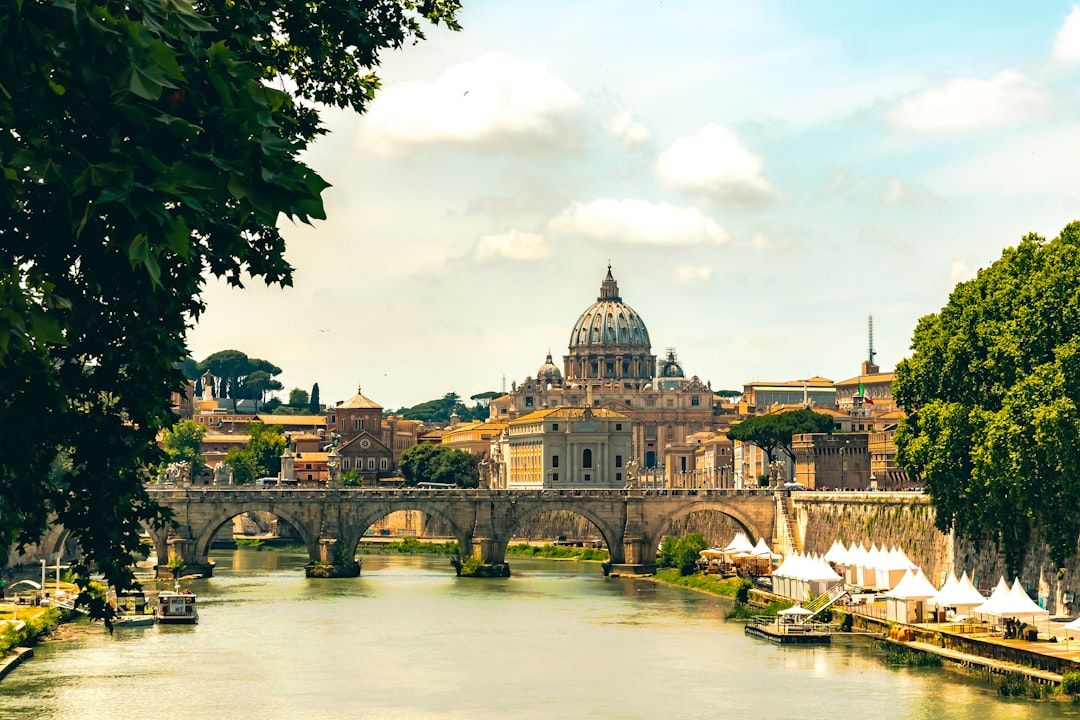 Riverside arched bridge to St Peters Basilica, reflective river surface mirrors classical dome, treelined embankments framing